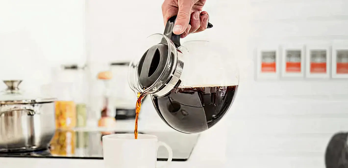 Person pouring coffee from a glass carafe into a white mug in a kitchen setting.