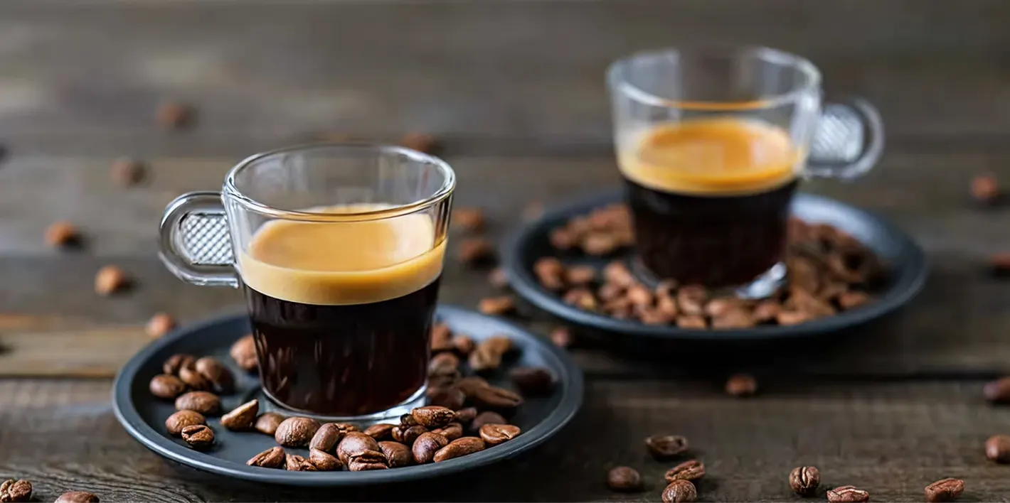 Two espresso cups on dark plates with coffee beans scattered around on a wooden surface.